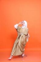 vol lengte studio portret van jong elegant gewoontjes professioneel moslim Indonesië vrouw met wit hijab in vervelend kaftan, hoog hakken zittend Aan stoel geïsoleerd Aan oranje achtergrond. foto