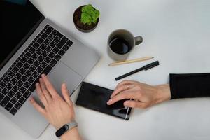 bovenaanzicht witte bureautafel met blanco notebook, computerlaptoptoetsenbord, pen, plant en andere kantoorbenodigdheden. koffiekopje met kopieerruimte, plat gelegd. vrouw handen met telefoon foto