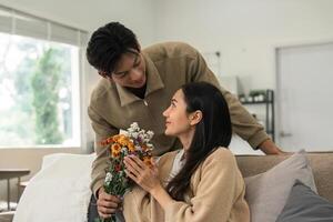 romantisch jong Aziatisch paar omarmen met Holding bloemen en glimlachen in leven kamer Bij huis. vallen in liefde. Valentijn concept foto