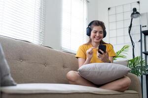 jong Aziatisch vrouw gebruik makend van mobiel telefoon en oortelefoons naar luisteren naar muziek- of online chatten Bij huis foto