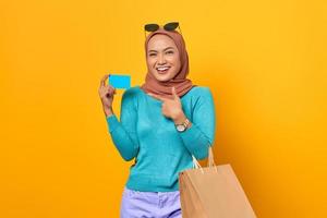 vrolijke jonge aziatische vrouw winkelen wijzende vinger naar creditcard op gele achtergrond foto