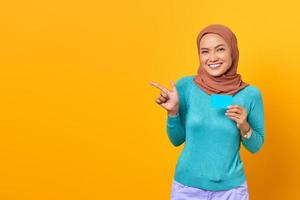 glimlachende jonge aziatische vrouw die met de vinger naar de kopieerruimte wijst en een creditcard op een gele achtergrond houdt