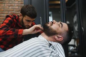 echt gebaard Mens krijgen baard kapsel met een Rechtdoor scheermes door kapper terwijl zittend in stoel Bij kapperszaak. kapperszaak thema. foto