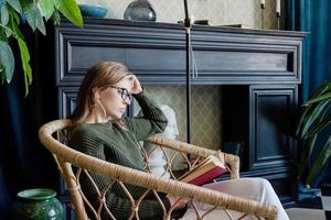 jonge blonde vrouw zit in een comfortabele stoel en leest een boek foto