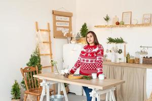 gelukkige jonge vrouw in kersttrui die warme dranken maakt in de keuken foto