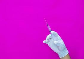 een gehandschoende hand op een roze achtergrond, met een medische spuit die wordt gebruikt om medicijnen en vitamines in het lichaam te injecteren. foto