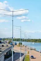 het ritme van hoge moderne lantaarnpalen aan de oever van de rivier tegen de achtergrond van een blauwe lucht met wolken.
