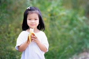 Aziatisch meisje zoete glimlach. kind met gele banaan in haar handen. kind dat fruit eet. kopieer ruimte natuur achtergrond. foto