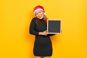 portret van glimlachende jonge aziatische vrouw in de hoed van de kerstman met schoolbord geïsoleerd over gele achtergrond foto