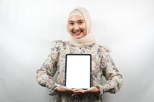 mooie jonge aziatische moslimvrouw die lacht, opgewonden en vrolijk tablet vasthoudt met wit of leeg scherm, app promoot, product promoot, iets presenteert, geïsoleerd op een witte achtergrond