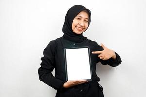 mooie jonge aziatische moslimvrouw die lacht, hand met tablet met wit of leeg scherm, app promoot, product promoot, iets presenteert, opgewonden en vrolijk, geïsoleerd op witte achtergrond