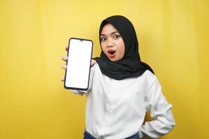 mooie jonge aziatische moslimvrouw geschokt, verrast, wauw-uitdrukking, hand met smartphone met wit of leeg scherm, app promoten, product promoten, iets presenteren, geïsoleerd foto