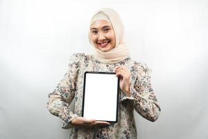mooie jonge aziatische moslimvrouw die lacht, opgewonden en vrolijk tablet vasthoudt met wit of leeg scherm, app promoot, product promoot, iets presenteert, geïsoleerd op een witte achtergrond