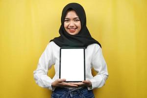 mooie jonge aziatische moslimvrouw glimlachend, opgewonden en vrolijk met tablet met wit of leeg scherm, app promoten, product promoten, iets presenteren, geïsoleerd op gele achtergrond