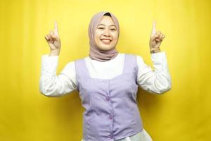 mooie jonge aziatische moslimvrouw die zelfverzekerd, enthousiast en vrolijk glimlacht met handen die omhoog wijzen en iets presenteren, kijkend naar camera geïsoleerd op gele achtergrond, reclameconcept