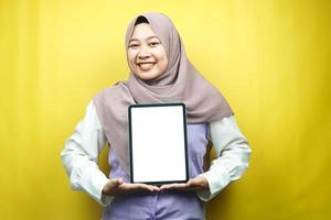mooie jonge aziatische moslimvrouw glimlachend, opgewonden en vrolijk met tablet met wit of leeg scherm, app promoten, product promoten, iets presenteren, geïsoleerd op gele achtergrond foto