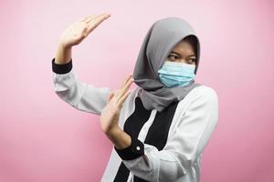 moslimvrouw die medisch masker draagt met hand die iets afwijst, hand die iets stopt, hand die iets niet leuk vindt, geïsoleerd op roze achtergrond