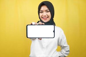 mooie jonge aziatische moslimvrouw die zelfverzekerd, enthousiast en vrolijk glimlacht met handen die smartphone vasthouden, toepassing promoten, spel promoten, geïsoleerd op gele achtergrond, reclameconcept