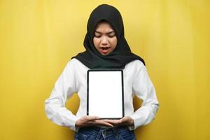 mooie jonge aziatische moslimvrouw glimlachend, opgewonden en vrolijk met tablet met wit of leeg scherm, app promoten, product promoten, iets presenteren, geïsoleerd op gele achtergrond