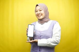 mooie jonge Aziatische moslimvrouw glimlachend vol vertrouwen en opgewonden met handen met smartphone, app promoten, iets promoten, geïsoleerd op gele achtergrond, reclameconcept