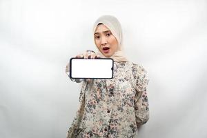 mooie jonge aziatische moslimvrouw geschokt, verrast, wauw-uitdrukking, hand met smartphone met wit of leeg scherm, app promoten, product promoten, iets presenteren, geïsoleerd foto