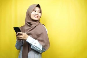 mooie jonge aziatische moslimvrouw glimlachend zelfverzekerd, enthousiast en vrolijk met de hand wijzend op smartphone, iets promoten, hand app promoten, geïsoleerd op gele achtergrond