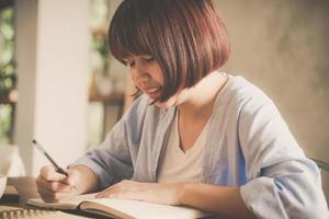 jonge zakenvrouw in witte jurk zittend aan tafel in café en schrijven in notitieblok. aziatische vrouw pratende smartphone en kopje koffie. freelancer werken in coffeeshop. foto