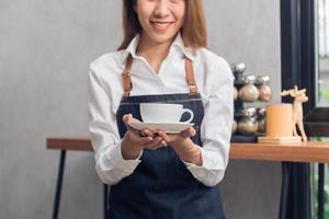 close-up van een jonge aziatische vrouwelijke barista die een kopje koffie vasthoudt aan haar klant met een glimlach omringd met een toogachtergrond. jonge vrouwelijke barista en haar kleine winkel. eten en drinken concept foto