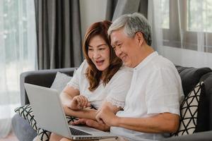 Aziatisch hoger paar dat laptop thuis met behulp van. Aziatische senior chinese grootouders, surfen op internet om sociale media te controleren terwijl ze op de bank liggen in het concept van de woonkamer thuis. foto