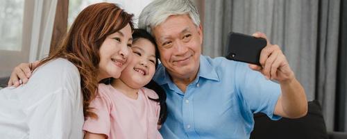 Aziatische grootouders selfie met kleindochter thuis. senior chinese gelukkige familie tijd doorbrengen ontspannen met behulp van mobiele telefoon met jong meisje kind liggend op de bank in de woonkamer. panoramische bannerachtergrond. foto