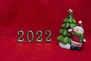 concept voor nieuwjaarskaart, uitnodiging en verpakking op een stoffen rode achtergrond een sneeuwpop met een kerstboom en gouden cijfers 2022 foto