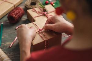 vrouw die kerst handgemaakte geschenkdoos maakt met bruin papier kromtrekken met kerst decor op houten tafel foto