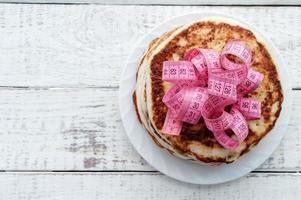 een stapel pannenkoeken op een bord met een centimeter tape erop op een houten tafel. het concept van een ongezonde levensstijl. overgewicht. foto