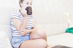 blonde met een kopje koffie zit thuis op de bank en werkt op een laptop. verre werk. werk vanuit huis. foto