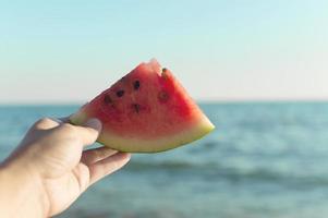 een hand houdt een plakje watermeloen vast tegen de achtergrond van de zee. het concept van toerisme en strandvakanties. foto