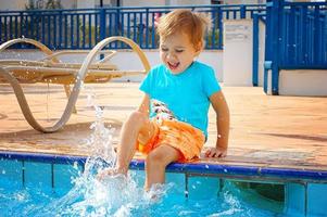 een kleine lachende jongen zit aan de rand van het zwembad, met zijn voeten in het water spetterend. toerisme, reizen. foto