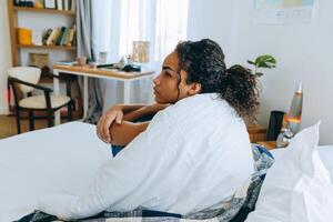 zijaanzicht van peinzende Afro-Amerikaanse vrouw wikkelde zichzelf in een deken op het bed foto