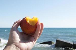 een hand houdt een perzik vast tegen de achtergrond van de zee. het concept van toerisme en strandvakanties. foto