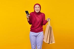 vrolijke jonge aziatische vrouw met mobiele telefoon en boodschappentassen op gele achtergrond foto