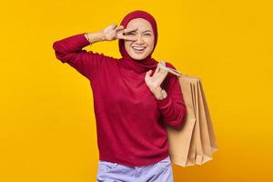 vrolijke jonge aziatische vrouw die vredesteken over ogen toont en boodschappentas op gele achtergrond houdt foto