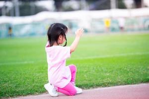 Kid ging zitten om zich voor te bereiden op haar run op het spoor van het stadion. kind draagt roze t-shirts. kinderen oefenen. zijaanzicht. foto