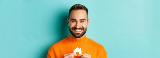 echt landgoed concept. gelukkig jong Mens zoeken voor huis huur, Holding huis papier maket en lachend, staand over- blauw achtergrond foto