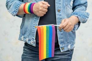 aziatische dame die een blauw spijkerjack of een denimoverhemd draagt en een regenboogvlag vasthoudt, symbool van de lgbt-trotsmaand viert jaarlijks in juni sociaal van homo, lesbienne, biseksueel, transgender, mensenrechten. foto
