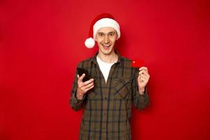 concept van mensen, online winkelen, technologie, kortingen. een opgewonden man in vrijetijdskleding kerst kerstman hoed maakt een bestelling via de telefoon betaalt creditcard, geïsoleerd op rode studio achtergrond foto