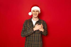 indoor shot van knappe man houdt handen op de borst, is hartelijk en vriendelijk, drukt dankbaarheid en dankbaarheid uit, heeft een aangename glimlach, gekleed in casual outfit kerstmuts, geïsoleerd op rode studiomuur foto