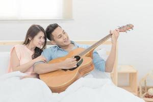 romantische huwelijksreis van Aziatische paarliefhebbers, man die gitaar speelt en zingt met zijn vrouw in de slaapkamer. foto