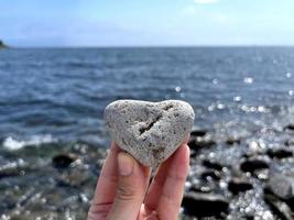 hartvorm steen in meisjes hand tegen de achtergrond van het strand. zomer zonnige dag. liefde, huwelijk en valentijnsdag concept. mooie en interessante stenen vinden. strandvakantie foto