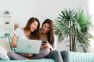 mooie jonge aziatische vrouwen lgbt lesbische gelukkige paar zittend op de bank online kopen met behulp van laptop een computer en telefoon in de woonkamer thuis. LGBT lesbisch koppel samen binnenshuis concept. foto