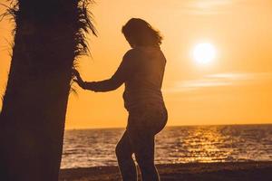 zwangere vrouw die haar buik aanraakt en naar de zonsondergang kijkt, naast een palmboom, in almerimar, almeria, spanje foto