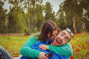 jong koppel zittend op het gras in een veld met rode papavers. het meisje kust de man terwijl hij een domme glimlach heeft. foto